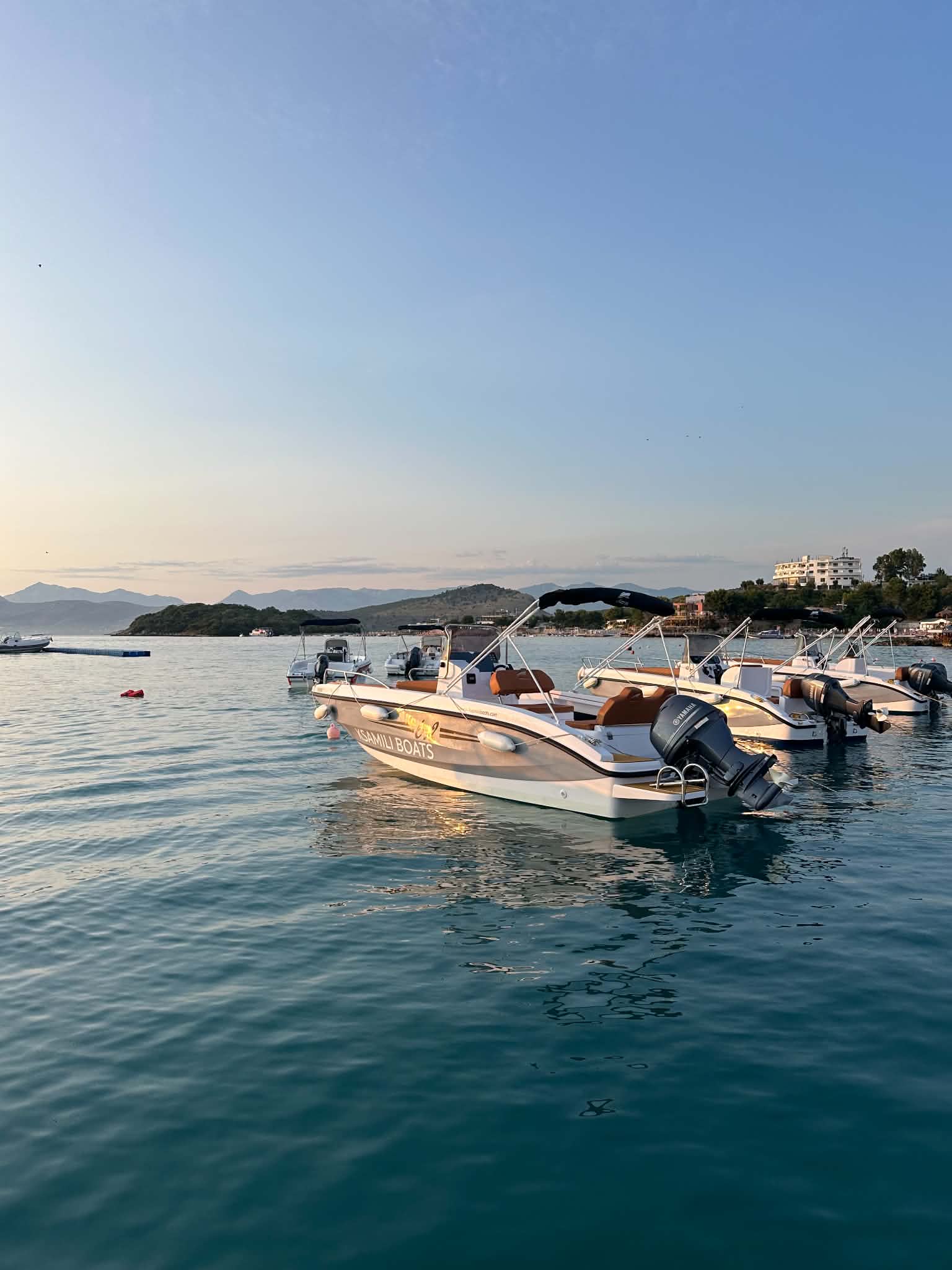 White Ksamili Boats bowrider moored in a row of open boats at golden hour, the Ksamil headland and hills behind